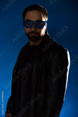 portrait of a man in a studio in a black leather coat. model with a beard on a blue background. trendy luminous glasses on the face