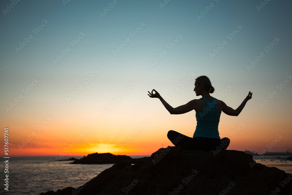 A woman doing yoga, meditating in the lotus pose on the beach during sunset.