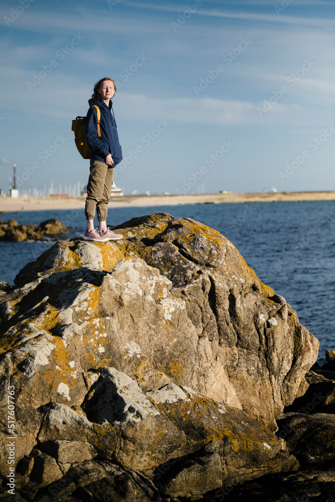 Obraz premium A woman tourist with a backpack stands on the sea coastal rocks.