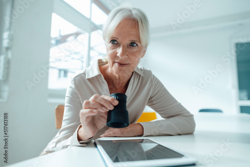 Senior businesswoman holding magnifying glass sitting with tablet PC in office photo