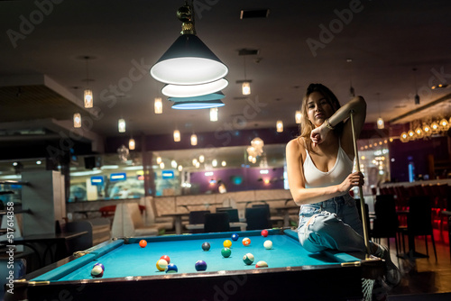 Φωτογραφία Glamorous brunette woman holding a cue and sitting on the billiard table