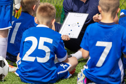 Sports Education For School Kids. Young Coach Explaining Game Tactics to Children Soccer Team. Kids in Blue Jersey Shirts Sitting With Young Coach and Planning Final Tournament Match Strategy