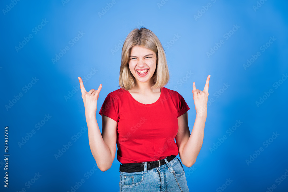Young and attractive caucasian blonde girl in casual clothes screaming and showing rocker and punk gesture isolated on blue studio background. Young and attractive caucasian blonde girl in casual clothes screaming and showing rocker and punk gesture isolated on blue studio background.