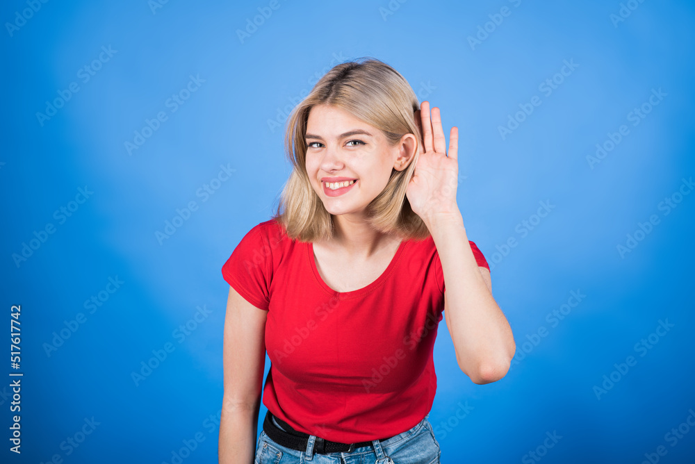 Fototapeta premium Young and attractive Caucasian blonde girl in casual clothes holding her hand to her ear and listening to the chatter isolated on a blue studio background.