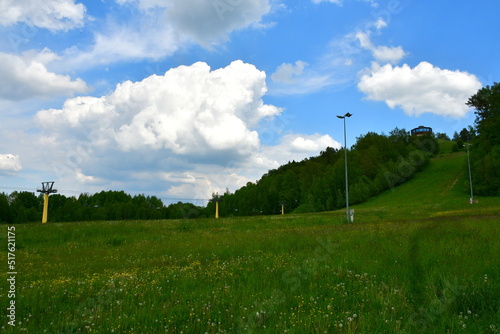A view of a ski lift with seats for passengers seen on a sunny summer day in the middle of a vast field, meadow, or pastureland, next to a tall hill or mountain used by tourists and skiers in Poland