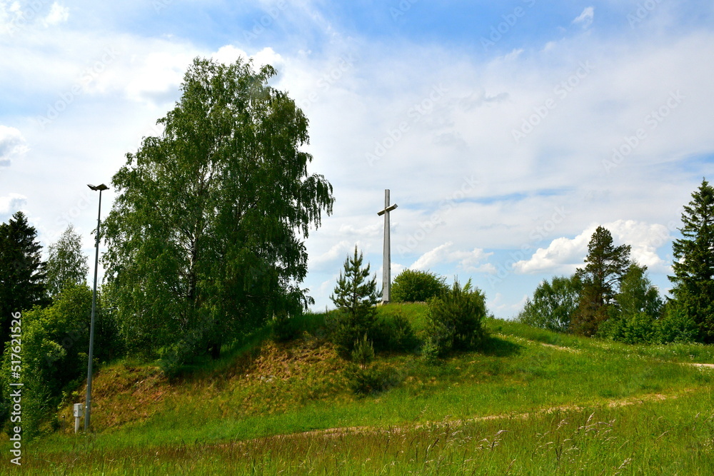 A view of a tall metal cross standing on top of a tall hill next to some tall trees, shrubs, flora, and other greenery seen on a sunny summer day on a Polish countryside during a hike