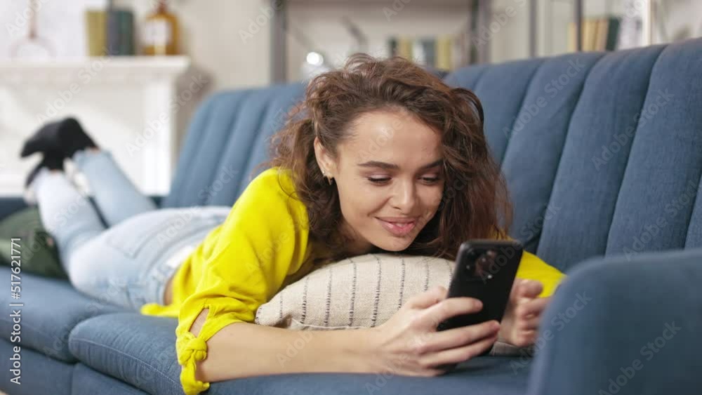 Attractive young woman lying on comfy sofa with modern smartphone in hands. Smiling female brunette scrolling social network or chatting online while resting at home. Attractive young woman lying on comfy sofa with modern smartphone in hands. Smiling female brunette scrolling social network or chatting online while resting at home.