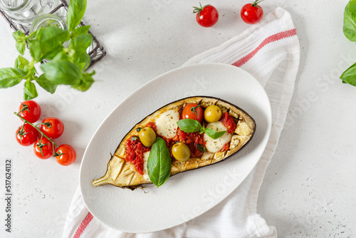 Italian traditional dish with eggplant, tomatoes, olives, mozzarella and basil. Baked aubergine stuffed with vegetables on a white plate. Vegan menu recipe. Top view.