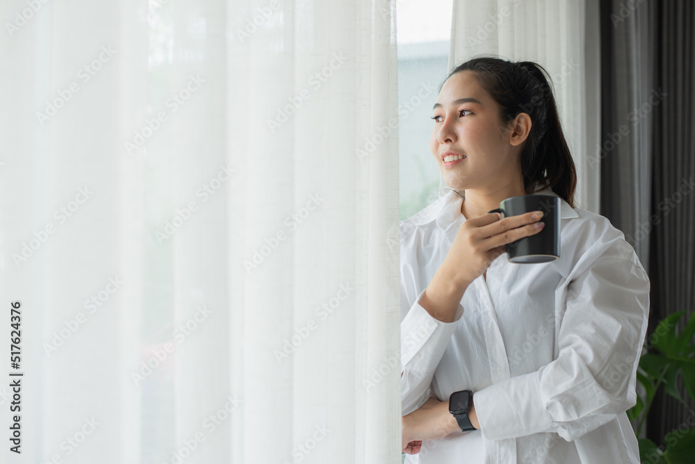 Young woman wearing white shirt smiling and standing by the window holding a cup of coffee in the morning. She likes to wake up to coffee in the morning, it makes her feel refreshed and happy. Young woman wearing white shirt smiling and standing by the window holding a cup of coffee in the morning. She likes to wake up to coffee in the morning, it makes her feel refreshed and happy.