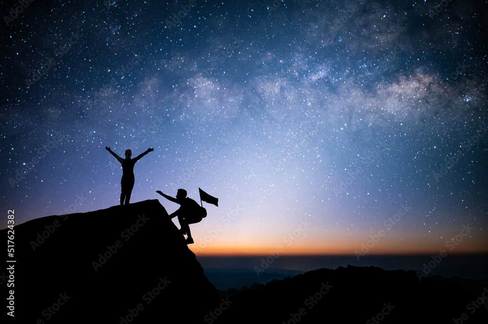 Two young tourist with backpack is climbing a cliff with beautiful night sky, Milky Way and star over the sky. He had an effort to climb all the way to the top of the mountain and he had to succeed.