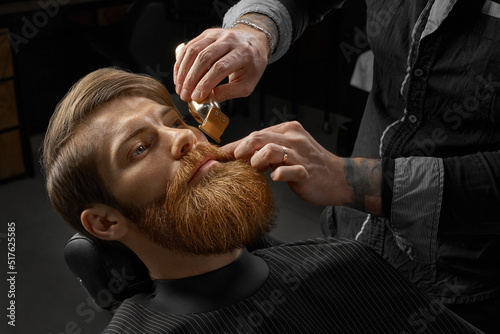 Fototapeta samoprzylepna Barbershop Shaving. Male barber shaves beard. Close-up of young bearded man getting beard haircut by hairdresser or barber at barbershop.
