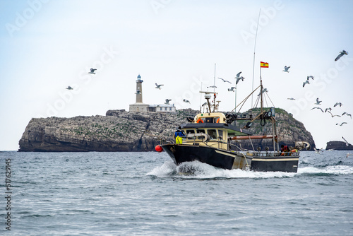 Barco pesquero volviendo a puerto rodeado de gaviotas frente a isla con faro