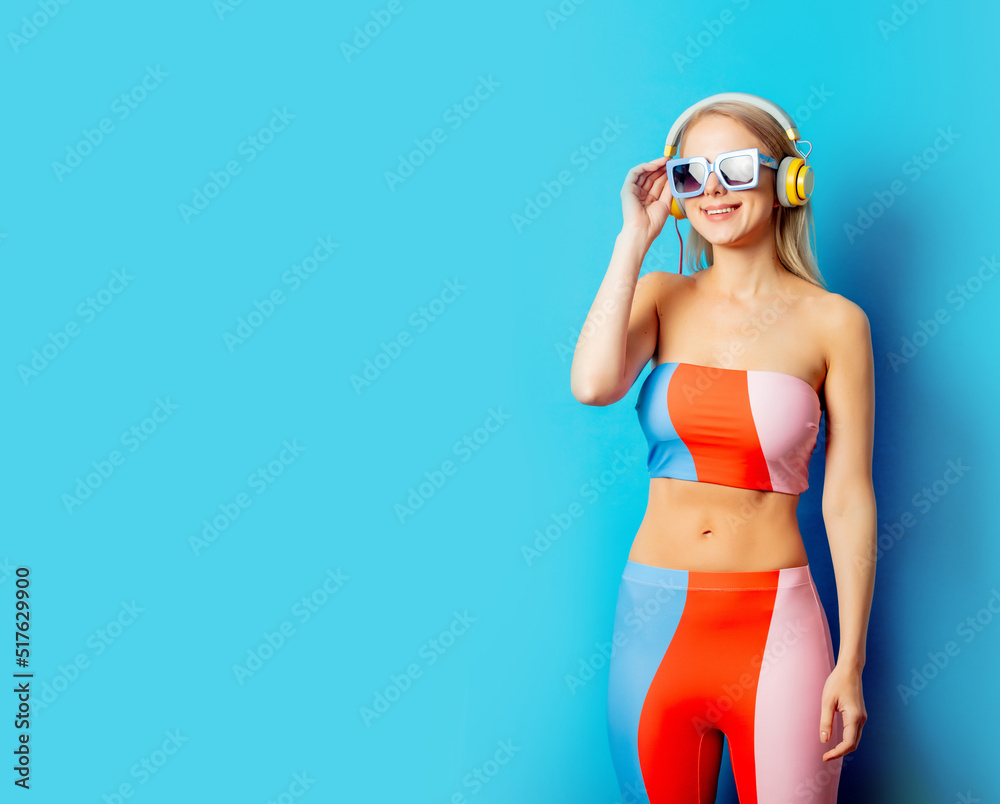 Stylish blond woman in colored swimming suit with headphones and sunglasses on blue background