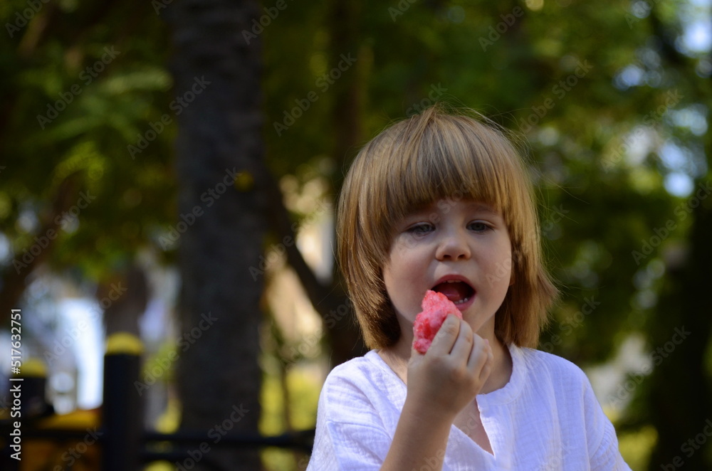 A little boy eats a pink cotton candy. Close-up of a child's face. Palm trees in the background. Concept: summer mood, holidays, travel, vacation. A little boy eats a pink cotton candy. Close-up of a child's face. Palm trees in the background. Concept: summer mood, holidays, travel, vacation.