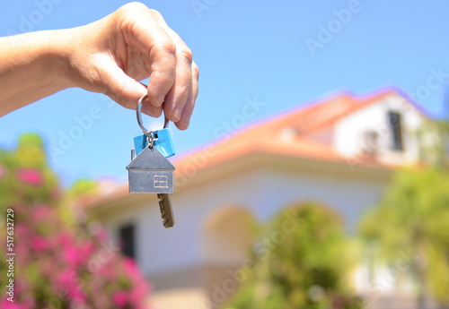 A hand holds a key against the backdrop of a villa with a garden. Concept: real estate transaction, buying a villa, own house.