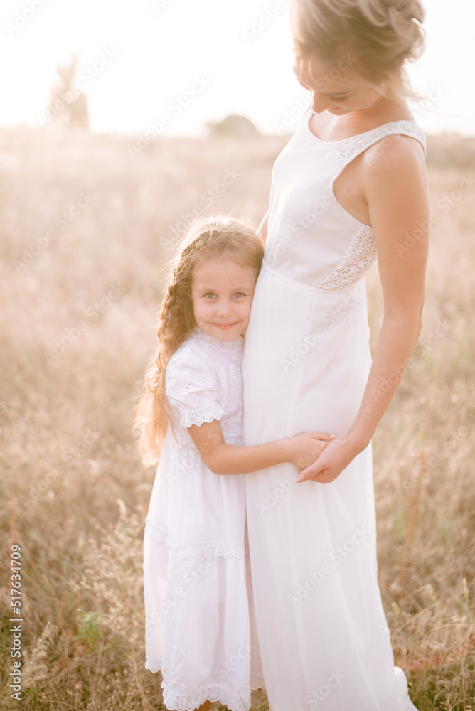 Fototapeta premium A cute little girl with long blond curly hair and her mother in a white summer dress and a straw boater hat in a field in the countryside in summer at sunset. Nature and Ecolife