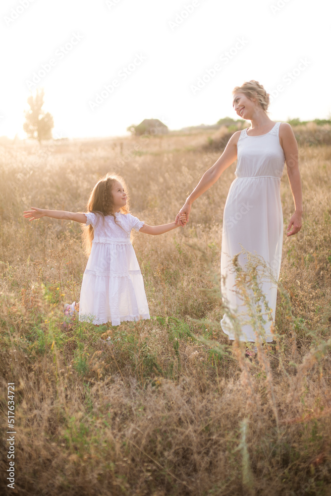 Fototapeta premium A cute little girl with long blond curly hair and her mother in a white summer dress and a straw boater hat in a field in the countryside in summer at sunset. Nature and Ecolife