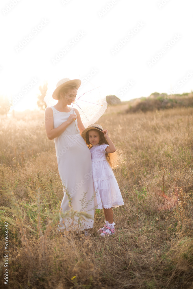 Fototapeta premium A cute little girl with long blond curly hair and her mother in a white summer dress and a straw boater hat in a field in the countryside in summer at sunset. Nature and Ecolife
