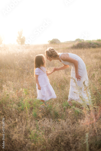 A cute little girl with long blond curly hair and her mother in a white summer dress and a straw boater hat in a field in the countryside in summer at sunset. Nature and Ecolife