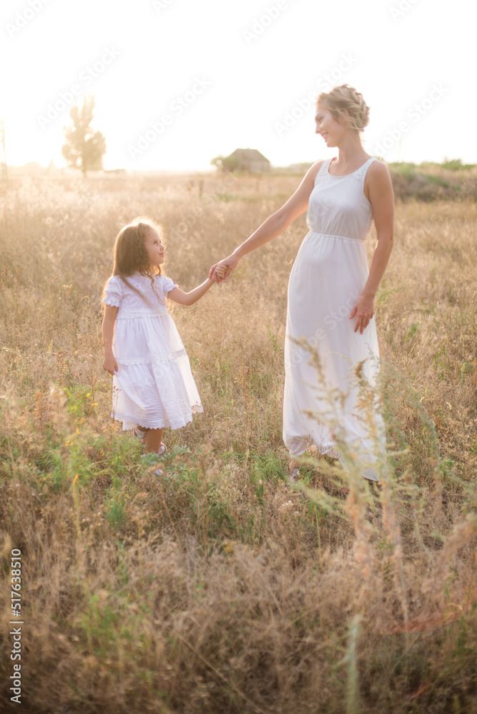 A cute little girl with long blond curly hair and her mother in a white summer dress and a straw boater hat in a field in the countryside in summer at sunset. Nature and Ecolife A cute little girl with long blond curly hair and her mother in a white summer dress and a straw boater hat in a field in the countryside in summer at sunset. Nature and Ecolife