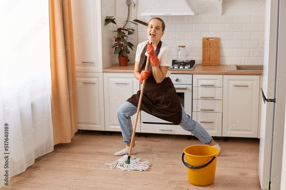 Full length photo of positive cheerful woman washing floor imagine she real celebrity holding mop, singing song, wear latex gloves and brown apron, posing in house indoors. Full length photo of positive cheerful woman washing floor imagine she real celebrity holding mop, singing song, wear latex gloves and brown apron, posing in house indoors.