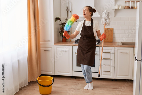 Indoor shot of happy positive woman wearing brown apron and jeans, doing the work about the house, holding mo in hand, using pp duster as a microphone, singing while cleaning her kitchen.