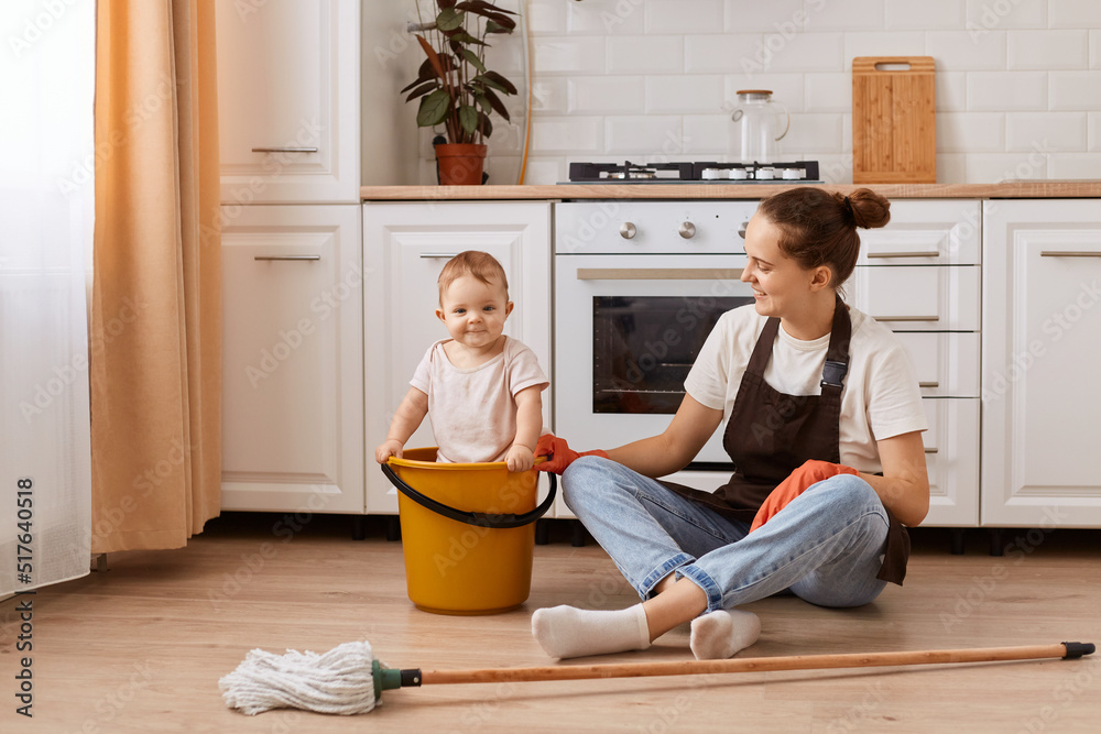 Indoor shot of attractive young adult female wearing brown apron sitting on floor after washing it and playing with her baby daughter in yellow bucket, mother looking at he child. Indoor shot of attractive young adult female wearing brown apron sitting on floor after washing it and playing with her baby daughter in yellow bucket, mother looking at he child.