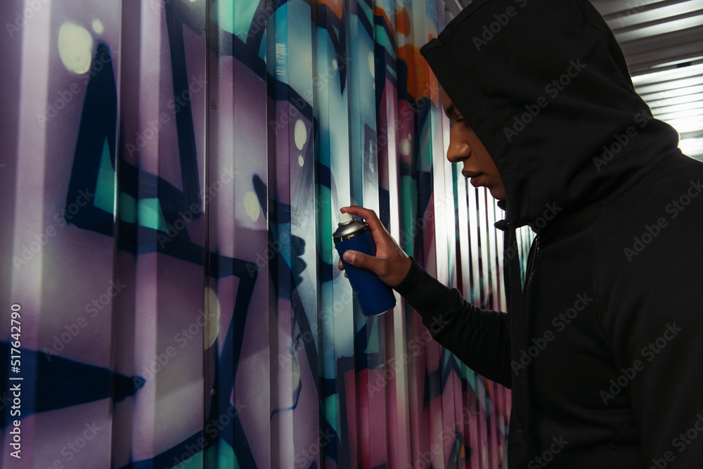 Side view of african american vandal in hood drawing graffiti on wall ...