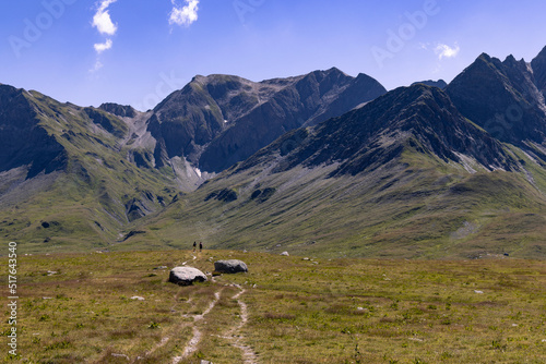 Two women walk on a winding alpine trail between mountains in late summer on a sunny day in the Greina Plateau, in Switzerland.