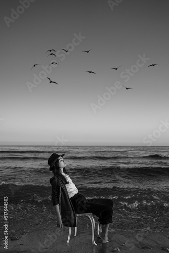 girl in a formal elegant stylish suit, shirt and hat sitting on a chair in the waves of the sea. artistic dreamer portrait of Creative black and white composition. birds in the sky. calm before storm photo