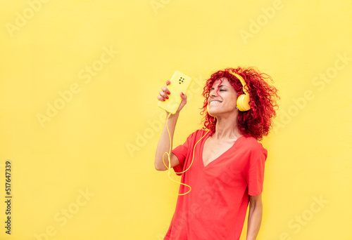 horizontal portrait of a latin woman with red afro hair holding a cell phone and enjoying listening to music with yellow headphones, dancing on a yellow background