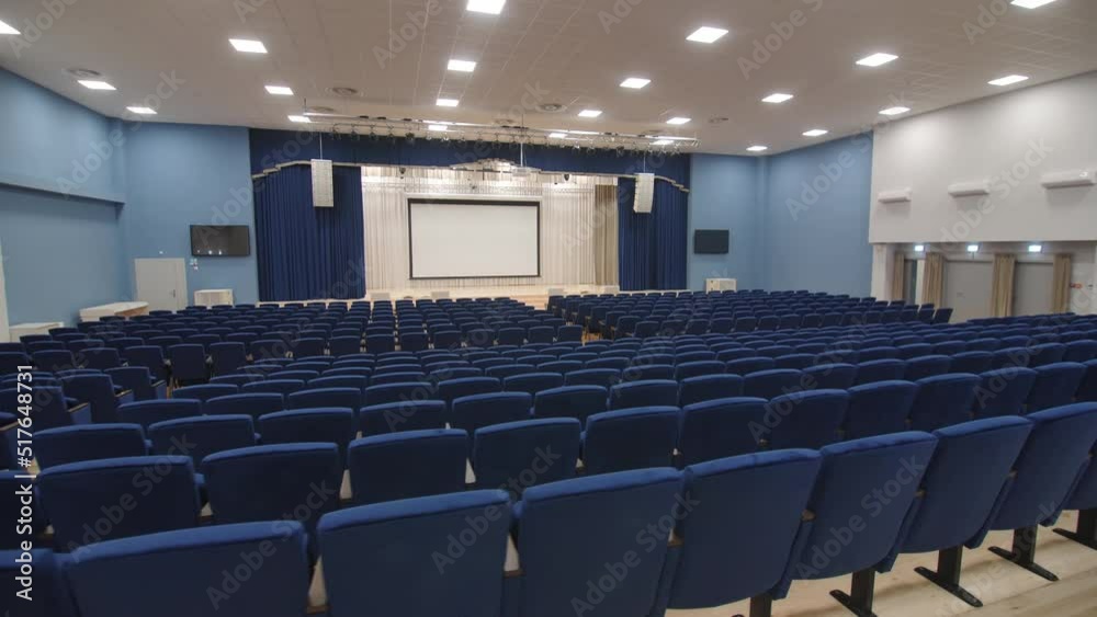 Large projection screen on stage and rows of soft chairs in empty spacious conference hall in business center
