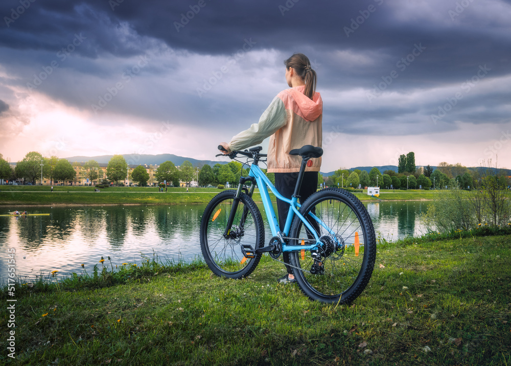 Woman riding a mountain bike near lake at overcast sunset in spring. Colorful landscape with sporty girl, bicycle, coast of river, green grass, cloudy sky in park in summer. Sport and travel. Biking Woman riding a mountain bike near lake at overcast sunset in spring. Colorful landscape with sporty girl, bicycle, coast of river, green grass, cloudy sky in park in summer. Sport and travel. Biking