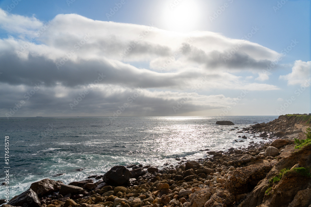 coastal shoreline with distant horizon, rocky beach and sunshine through a clouded blue sky