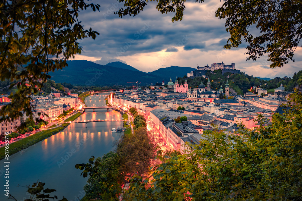Naklejka premium Illuminated streets in Salzburg town. Aerial summer view of Old City, birthplace of famed composer Mozart and Salzach river. Gloomy sunset in Eastern AlpsAustria, Europe. Traveling concept background.