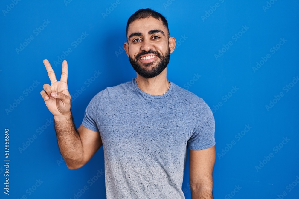 Middle east man with beard standing over blue background smiling with happy face winking at the camera doing victory sign with fingers. number two. Middle east man with beard standing over blue background smiling with happy face winking at the camera doing victory sign with fingers. number two.
