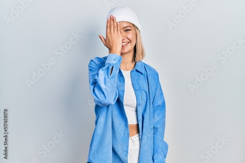 Beautiful blonde woman wearing wool hat covering one eye with hand, confident smile on face and surprise emotion.