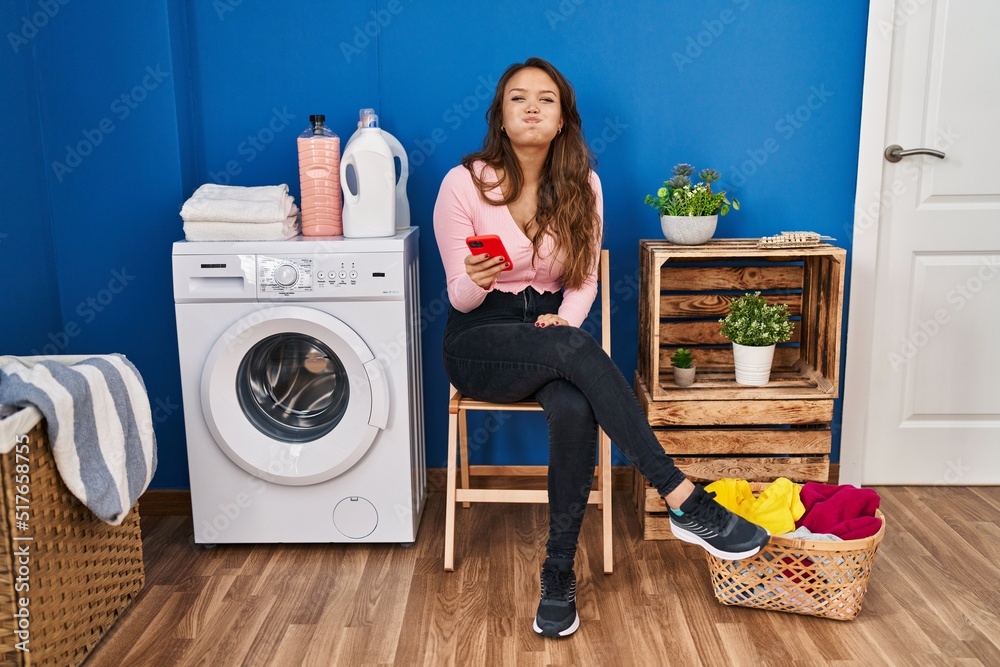 Young hispanic woman sitting waiting for laundry using smartphone puffing cheeks with funny face. mouth inflated with air, crazy expression. Young hispanic woman sitting waiting for laundry using smartphone puffing cheeks with funny face. mouth inflated with air, crazy expression.