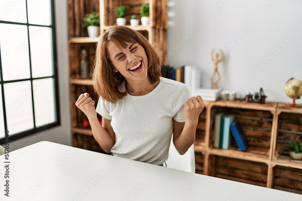 Young caucasian girl wearing casual clothes sitting on the table at home very happy and excited doing winner gesture with arms raised, smiling and screaming for success. celebration concept. Young caucasian girl wearing casual clothes sitting on the table at home very happy and excited doing winner gesture with arms raised, smiling and screaming for success. celebration concept.