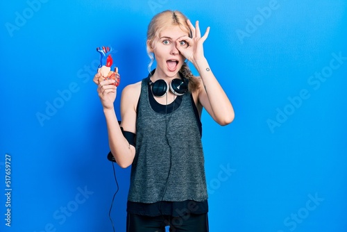 Beautiful caucasian sports woman with blond hair holding heart smiling happy doing ok sign with hand on eye looking through fingers photo