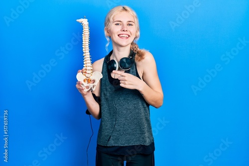 Beautiful caucasian sports woman with blond hair holding anatomical model of spinal column smiling happy pointing with hand and finger