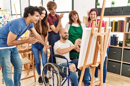 Group of young paint students smiling happy and looking draw of disabled partner sitting on wheelchair at art studio.