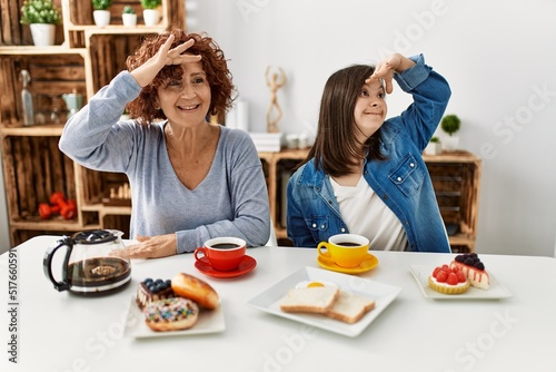 Family of mother and down syndrome daughter sitting at home eating breakfast very happy and smiling looking far away with hand over head. searching concept.