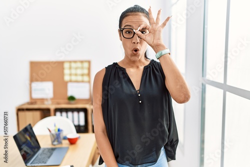 Young hispanic business woman working at the office doing ok gesture shocked with surprised face, eye looking through fingers. unbelieving expression.