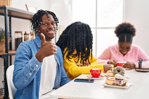Group of three young black people sitting on a table having coffee smiling happy and positive, thumb up doing excellent and approval sign