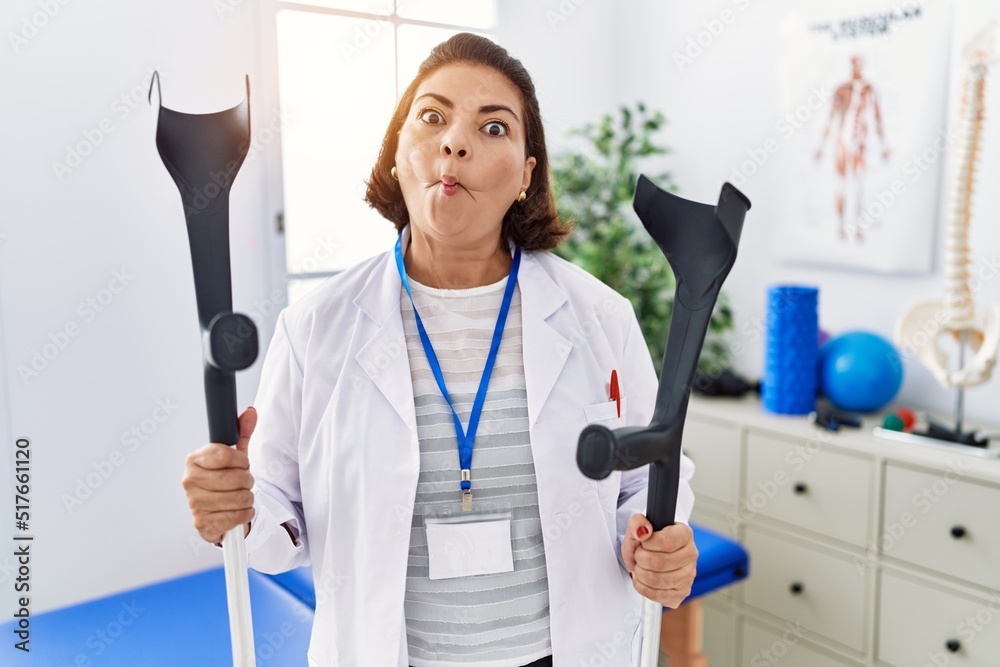Middle age hispanic physiotherapy woman holding crutches making fish face with mouth and squinting eyes, crazy and comical. Middle age hispanic physiotherapy woman holding crutches making fish face with mouth and squinting eyes, crazy and comical.