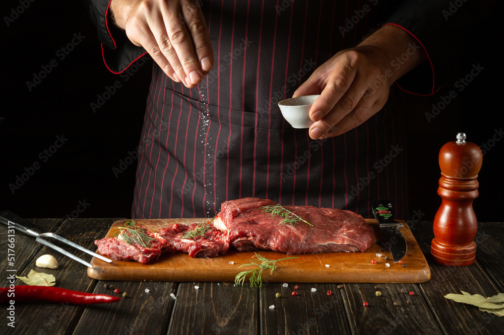 An experienced chef salts raw fresh beef before roasting or barbecuing. Working environment in the kitchen of a restaurant or hotel.