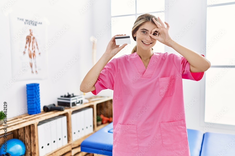 Young physiotherapist woman working at pain recovery clinic holding credit card smiling happy doing ok sign with hand on eye looking through fingers Young physiotherapist woman working at pain recovery clinic holding credit card smiling happy doing ok sign with hand on eye looking through fingers
