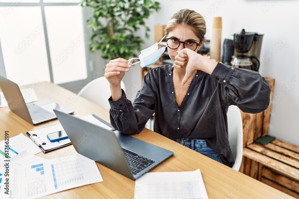 Young blonde woman working at the office wearing safety mask with angry face, negative sign showing dislike with thumbs down, rejection concept Young blonde woman working at the office wearing safety mask with angry face, negative sign showing dislike with thumbs down, rejection concept