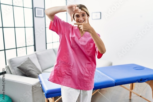 Young blonde woman working as physiotherapist at home smiling making frame with hands and fingers with happy face. creativity and photography concept.
